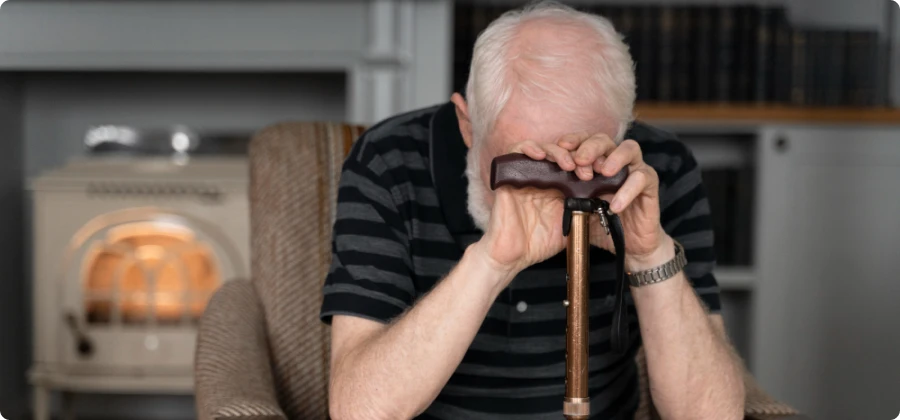 An elderly man sitting in a chair with his head bowed, holding a walking cane. He appears to be feeling overwhelmed or distressed in a cozy indoor setting.